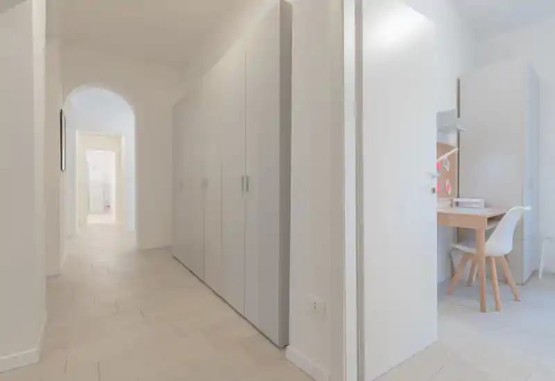 Bright, minimalist entrance hallway with built-in wardrobes and a tiled floor; a doorway on the right reveals a small workspace. Clean neutral tones create an airy first impression.
