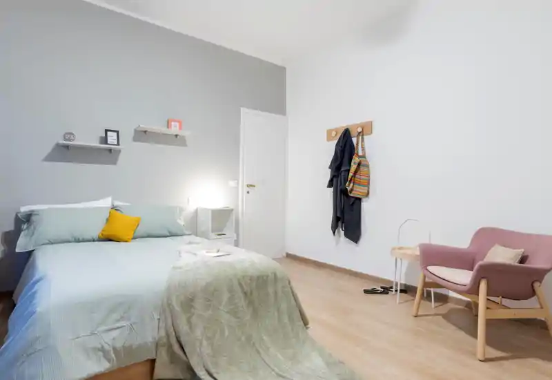 Camera da letto moderna e luminosa con letto matrimoniale rifatto, biancheria verde chiaro e una coperta accogliente. L'ambiente presenta mensole minimali sopra il letto, un comodino con lampada e una poltrona vicino ai ganci a muro.