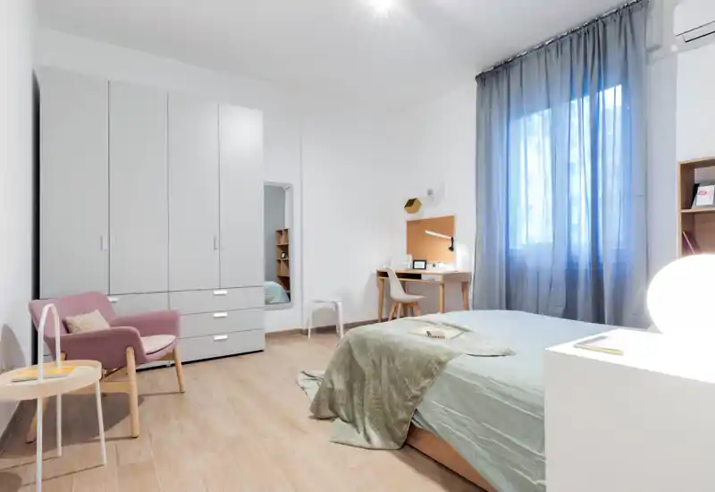 Camera da letto moderna e luminosa con letto matrimoniale ben rifatto, tessuti verdi, scrivania vicino alla finestra, armadio e una poltrona accogliente — perfetta per un soggiorno confortevole.