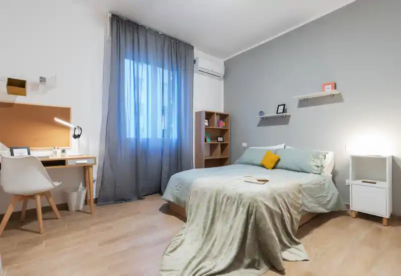 Camera da letto luminosa e moderna con letto matrimoniale, biancheria soffice e un plaid accogliente; una scrivania vicino alla finestra e mensole di design rendono lo spazio funzionale e curato.
