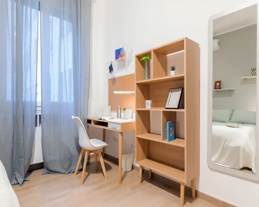 Angolo camera luminoso e ordinato con scrittoio, libreria e uno scorcio del letto riflesso nello specchio — ideale per uno studente o single.