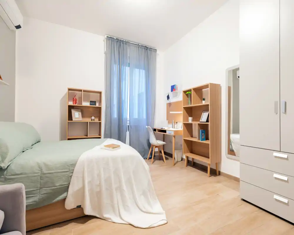 Camera da letto luminosa e moderna con letto ben rifatto, tessili morbidi e una scrivania compatta — ideale per un soggiorno singolo confortevole.