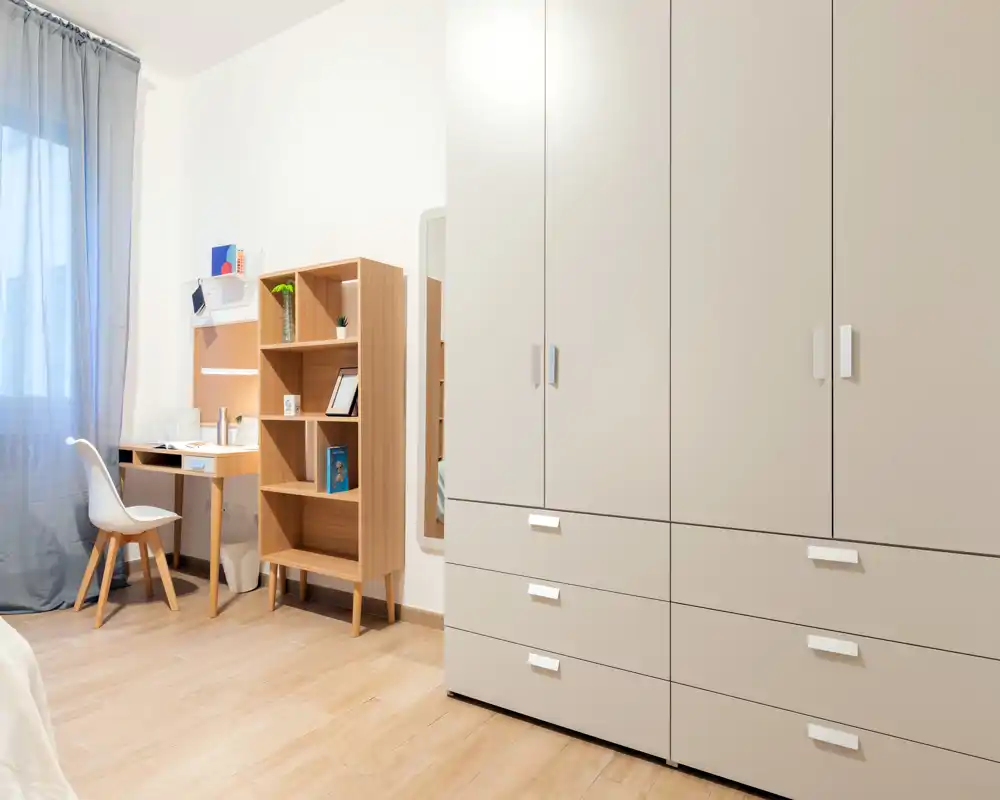 Angolo di camera luminoso e moderno con un grande armadio, scrivania e scaffalatura; una piccola porzione del letto è visibile sul bordo dell’immagine.