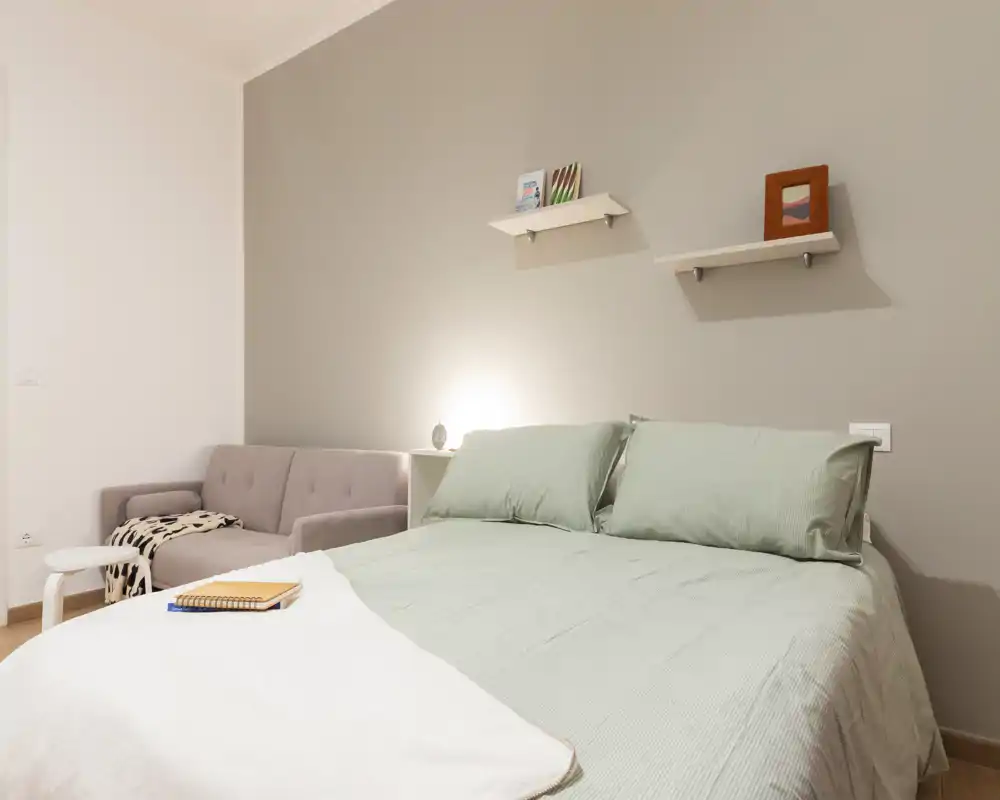 Camera da letto luminosa e ordinata con un letto matrimoniale ben rifatto in lenzuola verde chiaro e un piccolo divano nell'angolo, con mensole minimal e luce calda sul comodino.