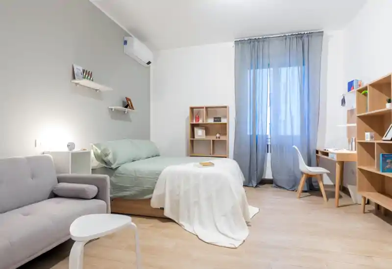 Camera da letto luminosa e moderna con letto matrimoniale ben rifatto, tessuti soffici e un piccolo angolo conversazione. Mobili in legno chiaro e scrivania vicino alla finestra creano un ambiente funzionale e tranquillo.