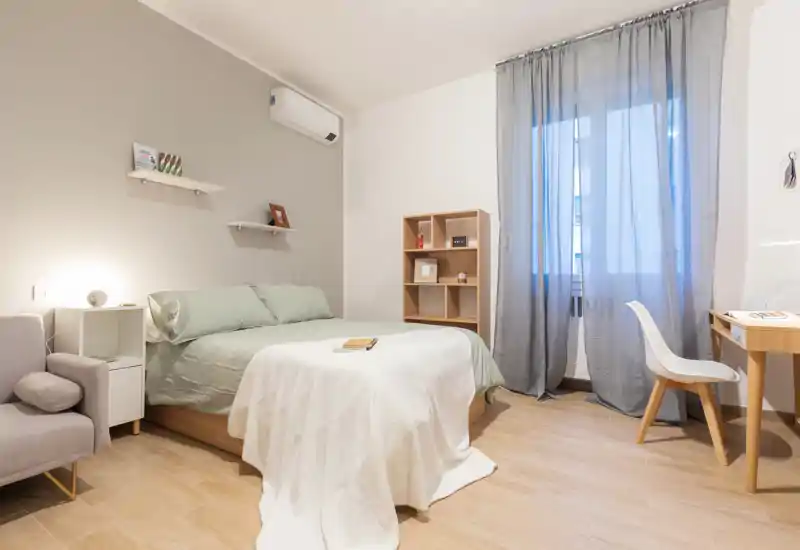 Camera da letto luminosa e ben arredata con letto matrimoniale, poltrona confortevole e una piccola scrivania vicino alla finestra. La luce naturale che filtra dalle tende crea un'atmosfera calda e accogliente, ideale per gli ospiti.