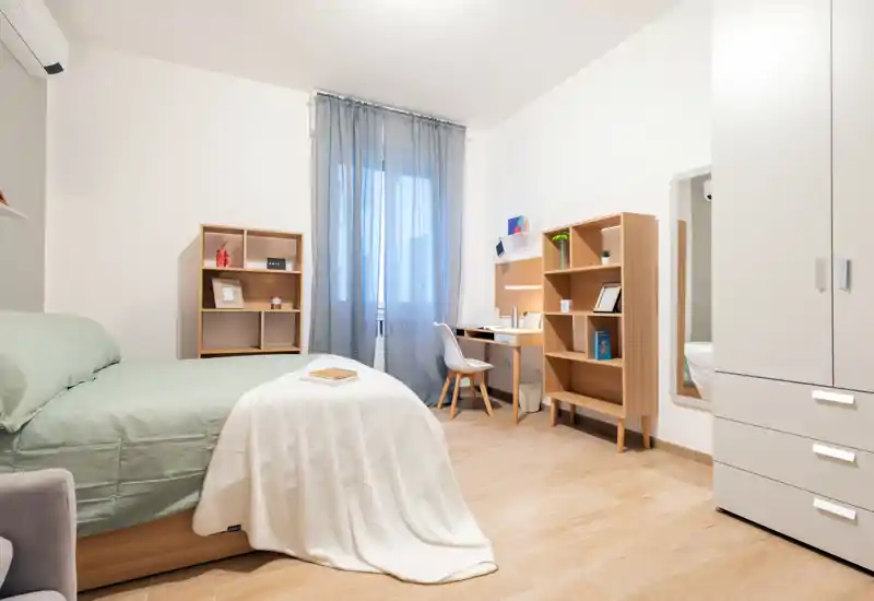 Camera da letto luminosa e moderna con letto ben rifatto, tessili morbidi e una scrivania compatta — ideale per un soggiorno singolo confortevole.