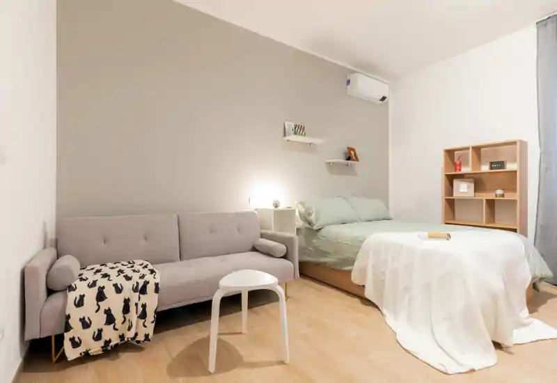 Camera da letto moderna e luminosa con letto matrimoniale, biancheria verde chiaro e un piccolo divano. Mensole minimal e una libreria a cubi in legno completano lo spazio caldo e ben illuminato, perfetto per annunci.