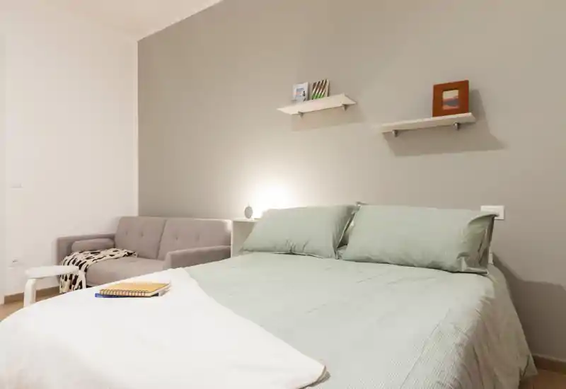 Camera da letto luminosa e ordinata con un letto matrimoniale ben rifatto in lenzuola verde chiaro e un piccolo divano nell'angolo, con mensole minimal e luce calda sul comodino.