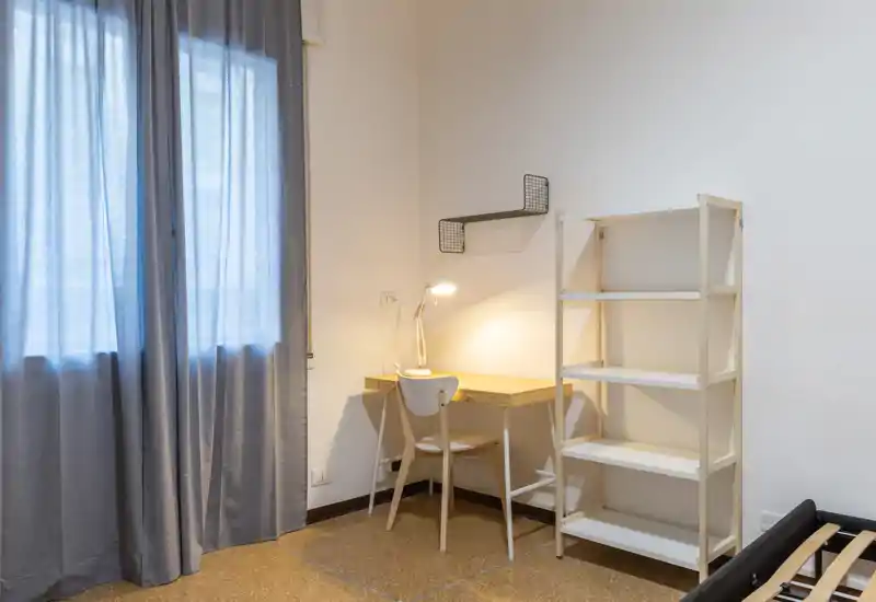 Angolo camera minimal con una piccola scrivania, sedia e scaffale; il telaio del letto è parzialmente visibile in basso a destra. Tonalità neutre, luce naturale soffusa e un angolo studio funzionale.