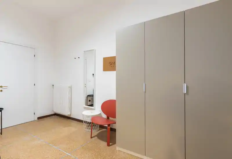 Ingresso luminoso e minimal con un grande armadio, radiatore, specchio e una piccola sedia rossa — spazio ordinato e funzionale per appendere giacche e riporre scarpe.
