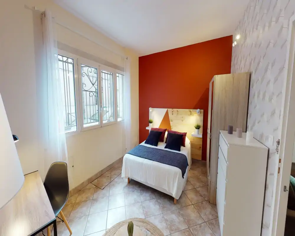 Camera da letto luminosa e curata con un comodo letto matrimoniale, cuscini decorativi e una parete d'accento rossa. La luce naturale entra da una grande finestra; armadio, cassettiera e scrivania completano l'ambiente.