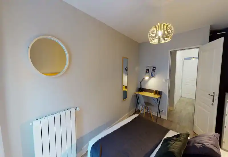 Chambre moderne et chaleureuse avec un lit double bien fait, coussins décoratifs et un petit bureau. Suspension lumineuse et miroir apportent du style, murs neutres pour une ambiance apaisante.