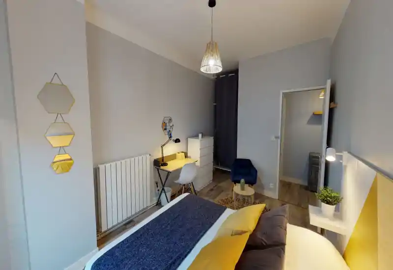 Camera da letto moderna e luminosa con un ampio letto in primo piano, cuscini giallo senape e plaid blu, scrivania compatta e un angolo salotto accogliente, perfetta per soggiorni comodi.