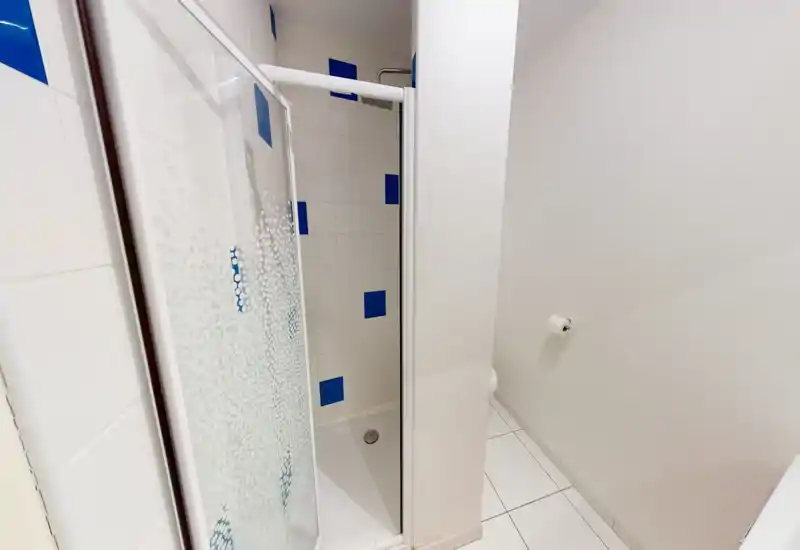 Angolo bagno compatto e pulito con cabina doccia piastrellata, inserti blu e porta scorrevole in vetro; pareti e pavimento bianchi per un aspetto luminoso e minimale.