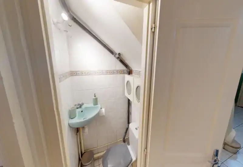 Bagno compatto con lavabo angolare e WC sotto un soffitto inclinato; pareti piastrellate bianche con bordo decorativo e impianto idraulico visibile, ben illuminato.