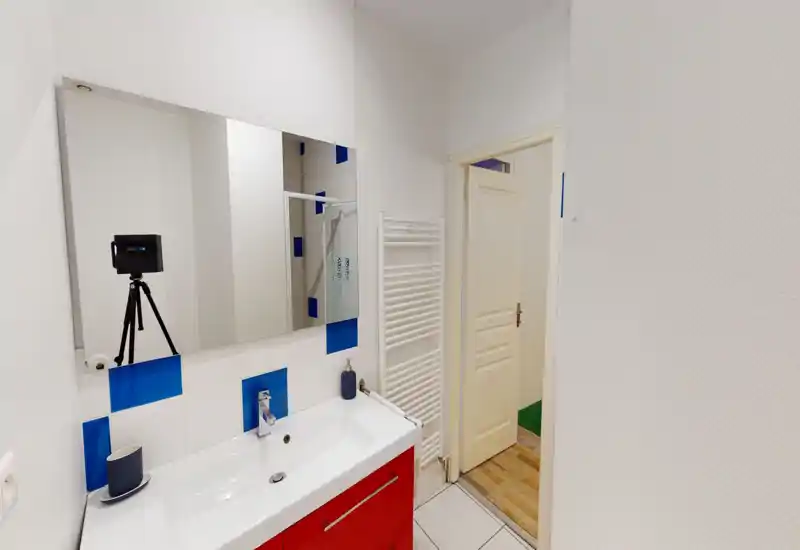 Bagno pulito e moderno con lavabo bianco, mobile rosso, grande specchio e piastrelle con dettagli blu. Spazio luminoso e minimal perfetto per foto dell'immobile.