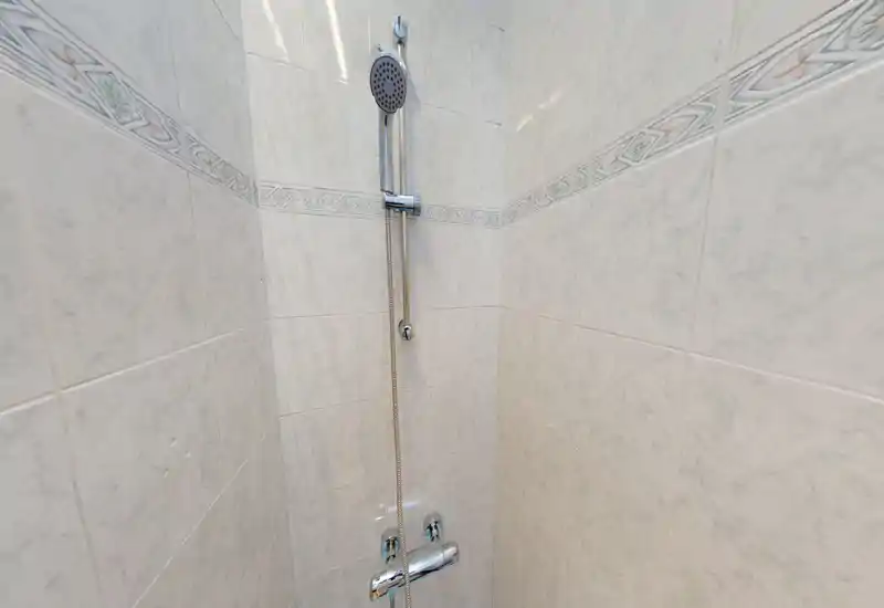 Doccia piastrellata compatta con soffione a mano cromato e rubinetteria; piastrelle pulite e fascia decorativa neutra.