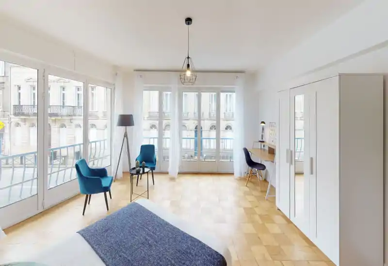 Chambre lumineuse et moderne avec de grandes fenêtres du sol au plafond, parquet en bois, lit bien fait au premier plan et coin salon avec deux fauteuils bleus. L'espace est aéré et élégant, avec un bureau et une armoire sur le mur droit.