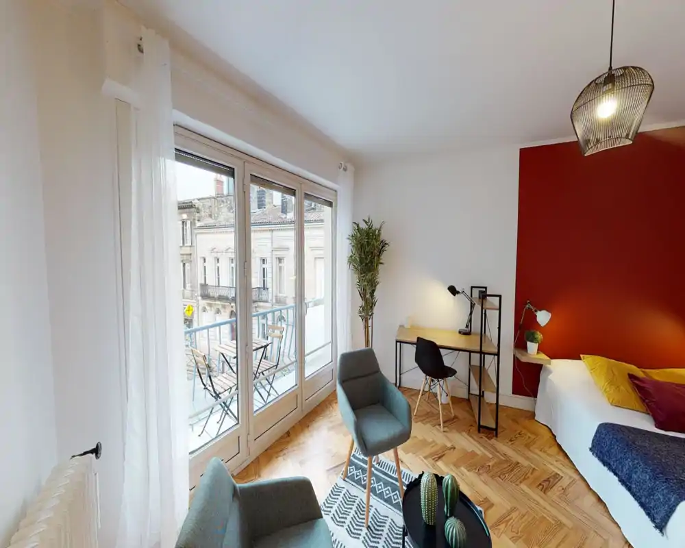 Dormitorio luminoso y moderno con pared en rojo, cama doble confortable, escritorio y grandes ventanales que dan a un pequeño balcón. Parquet de madera, textiles acogedores e iluminación moderna crean un ambiente cálido y atractivo para anuncios de alquiler.
