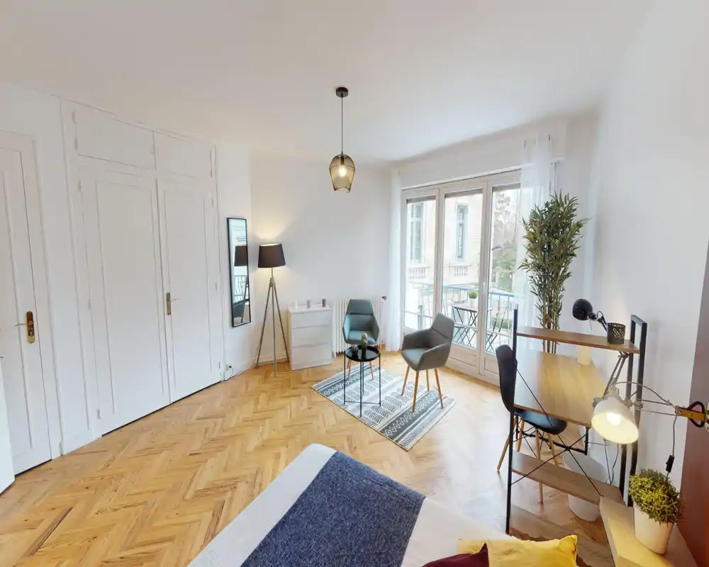 Bright, modern bedroom with wooden herringbone floors, a visible double bed in the foreground, a cozy seating area by large French windows and a small desk corner — ideal for a well-lit rental.