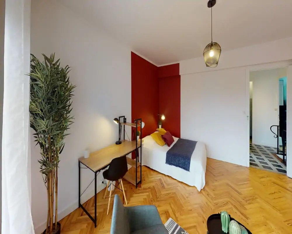 Bright, stylish bedroom with a comfortable single bed against a warm red accent wall, wooden parquet floors and a tidy workspace with desk and lamp — ideal for a student or short-stay guest.