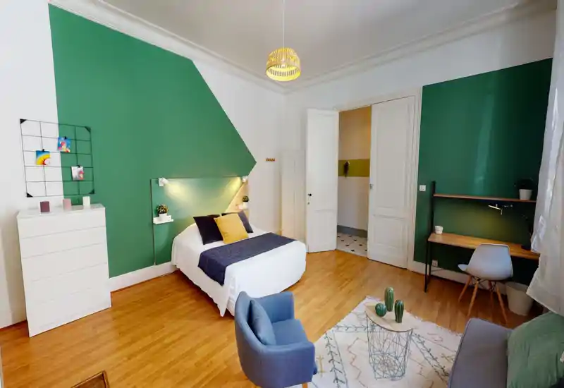 Camera da letto luminosa e di design con letto matrimoniale al centro, cuscini decorativi e un'area salotto accogliente. La parete verde, il pavimento in legno e l'angolo scrivania creano un'atmosfera moderna e invitante.