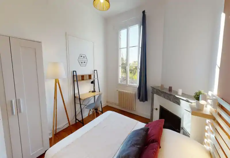 Chambre lumineuse et soignée avec lit double visible, parquet et un bureau près de la fenêtre. La lumière naturelle et les textiles apportent une atmosphère chaleureuse.