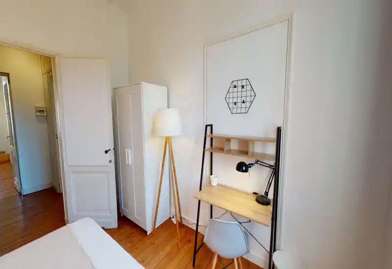 Coin chambre lumineux et minimaliste avec bord de lit visible, armoire, lampe sur trépied et petit bureau en bois avec lampe de travail — idéal pour un espace de travail calme.