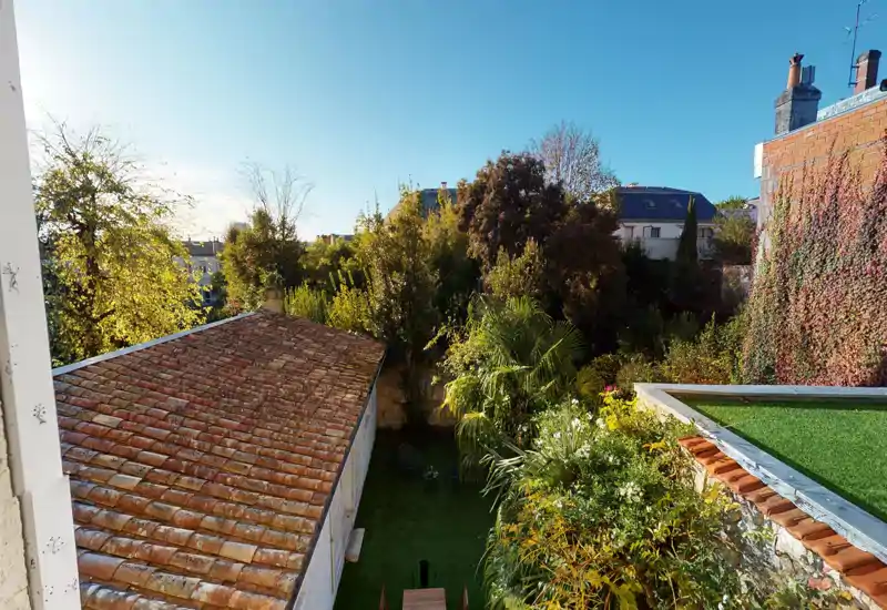 Vue lumineuse sur le jardin avec arbres matures, un toit en tuiles à gauche et une petite pelouse — photo extérieure idéale montrant la verdure et l'espace extérieur.