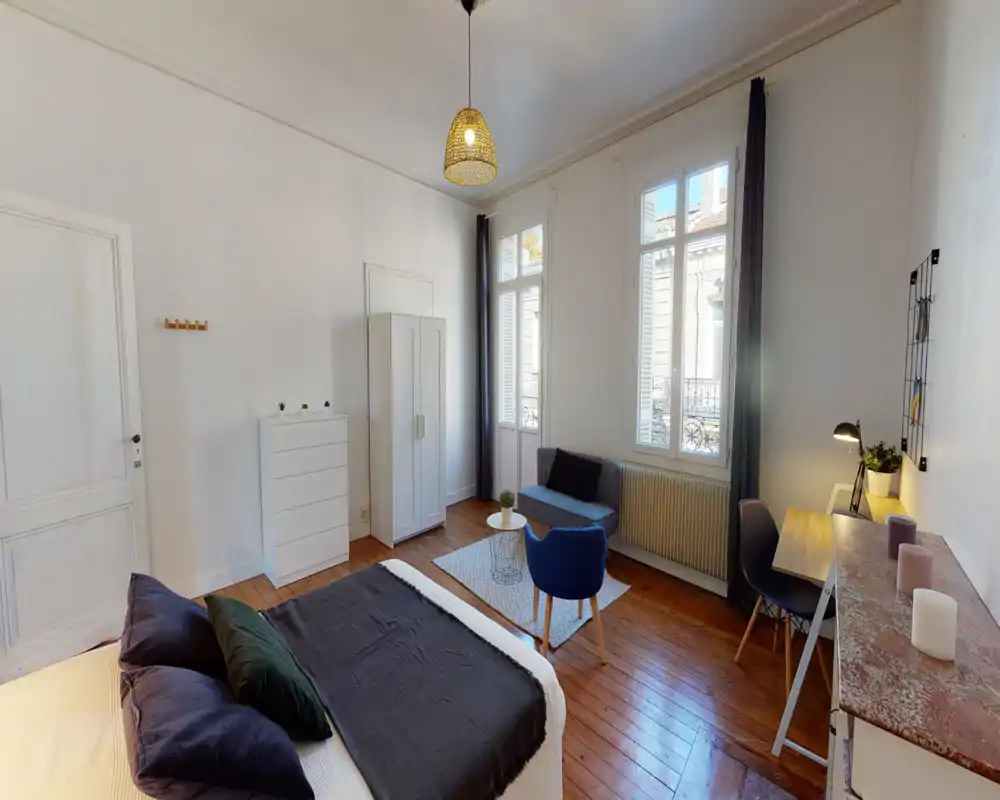 Camera da letto luminosa e curata: letto ampio in primo piano, pavimento in legno, area conversazione vicino alle finestre alte e una scrivania funzionale.