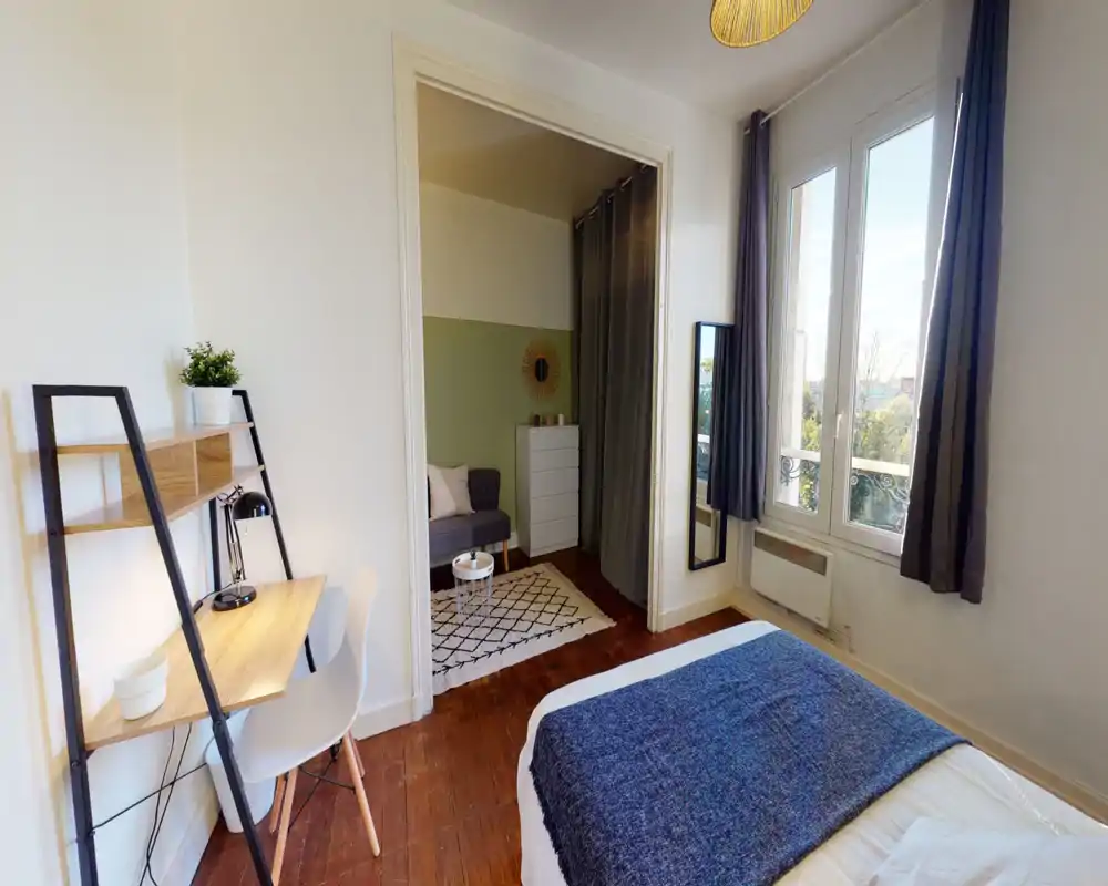 Spacious, bright bedroom with a comfortable double bed in the foreground, a desk workspace, large window with curtains and a small seating area separated by a curtained alcove.