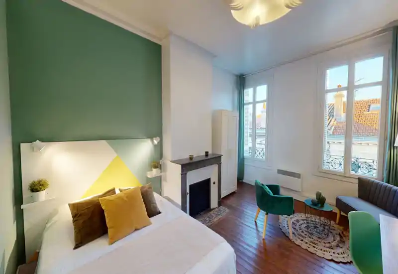 Chambre lumineuse et élégante avec un grand lit au premier plan, coussins décoratifs, mur d'accent vert et lumière naturelle par de grandes fenêtres. Coin salon confortable près de la fenêtre.