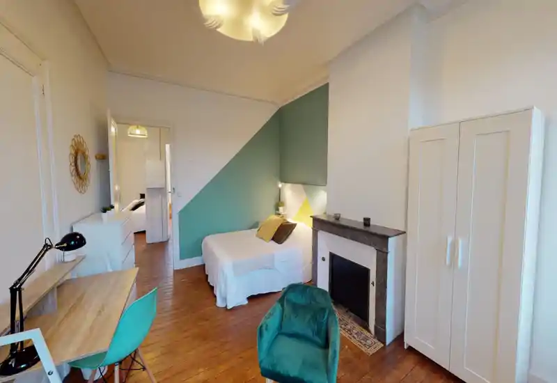 Chambre lumineuse et élégante avec un lit double contre un mur accentué vert-bleu, textiles confortables et décoration coordonnée ; comprend une armoire, un bureau et une cheminée décorative.