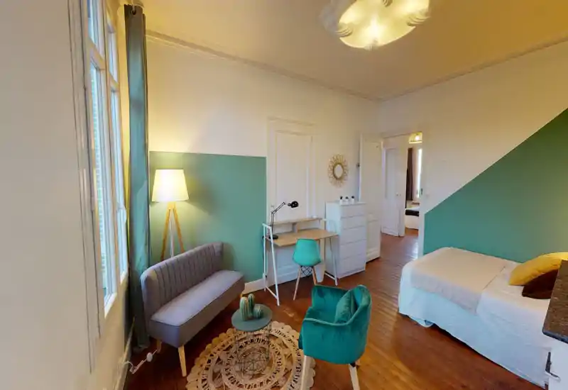 Chambre lumineuse et élégante avec un lit confortable, coussins décoratifs et un mur d'accent vert. Un coin salon, un bureau et la lumière naturelle offrent un espace polyvalent pour se détendre ou travailler.