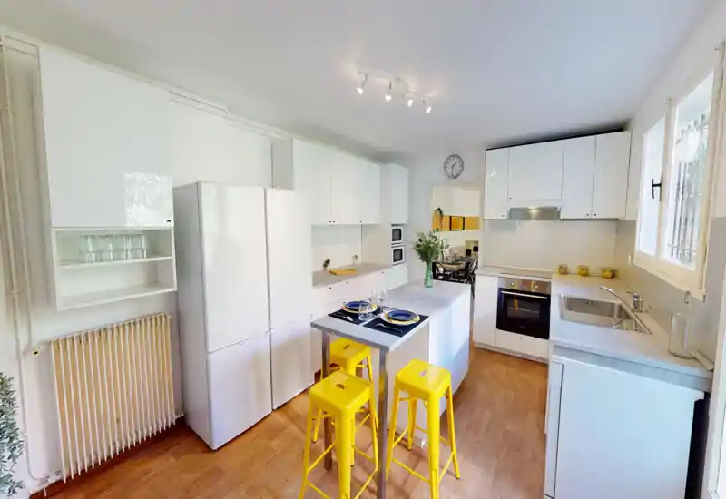 Cucina moderna e luminosa con mobili bianchi, isola apparecchiata per due e sgabelli gialli vivaci; finestra ampia e stile curato la rendono accogliente per cucinare e mangiare.