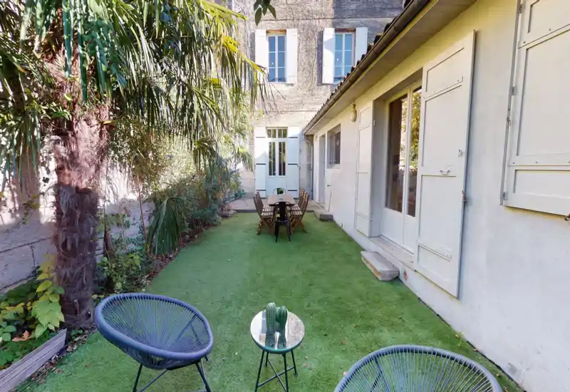 Cortile privato soleggiato con zona salotto e tavolo da pranzo su prato sintetico, bordato da palme e aiuole — ideale per pranzi rilassati e momenti conviviali.