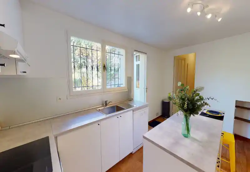 Cucina luminosa e ordinata con isola, lavabo sotto la finestra e vaso decorativo. Spazio ben illuminato con posti a sedere e piani di lavoro funzionali, ideale per un appartamento compatto.