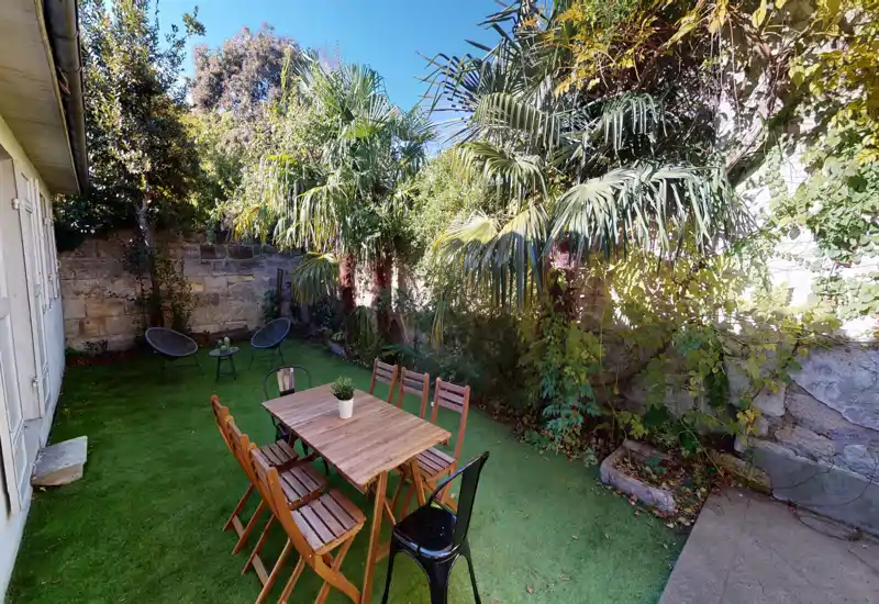 Giardino/terrazza privata soleggiata con tavolo da pranzo in legno e sedie su prato verde, circondata da palme e muri in pietra — perfetta per pranzi all'aperto e relax.