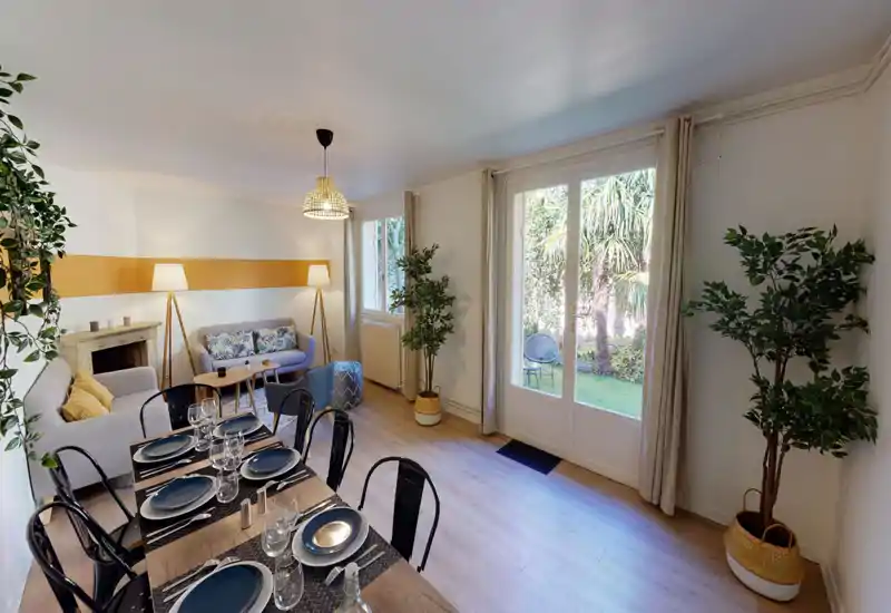 Soggiorno luminoso e curato con tavolo da pranzo in primo piano, divani confortevoli, cuscini decorativi, piante e una grande porta vetrata che si affaccia sul giardino.