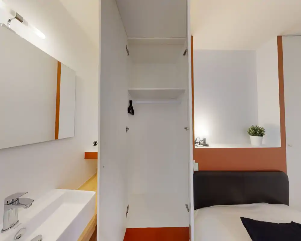 Chambre lumineuse et minimaliste montrant un lit bien fait avec tête de lit visible, une petite étagère de chevet avec lampe et plante, et une armoire ouverte. La photo met en valeur des lignes épurées et des touches de couleur chaleureuses.