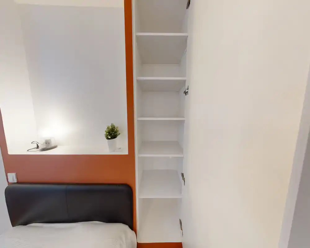 Vue compacte de la chambre montrant la tête de lit, une niche avec lampe et plante et des étagères ouvertes. Style moderne et épuré avec une touche de couleur chaude et un bon éclairage.