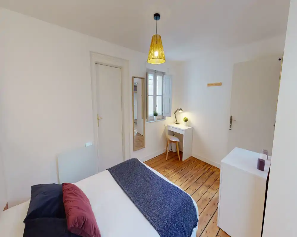 Chambre lumineuse et soignée avec parquet, lit double confortable et textiles superposés, petite table de travail près de la fenêtre — idéale pour travailler ou se détendre.