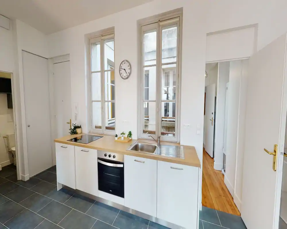 Cuisine moderne et lumineuse avec plan de travail en bois, four encastré et évier sous de grandes fenêtres ; meubles blancs et petites plantes pour une ambiance fonctionnelle et fraîche.