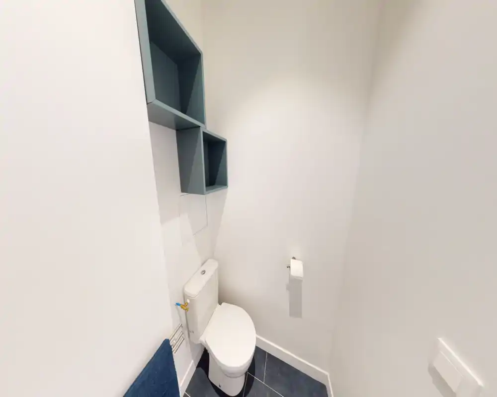 Petite salle de bains bien éclairée avec WC, étagères murales ouvertes et porte-papier. Murs blancs propres et carrelage sombre pour un style moderne et minimaliste.