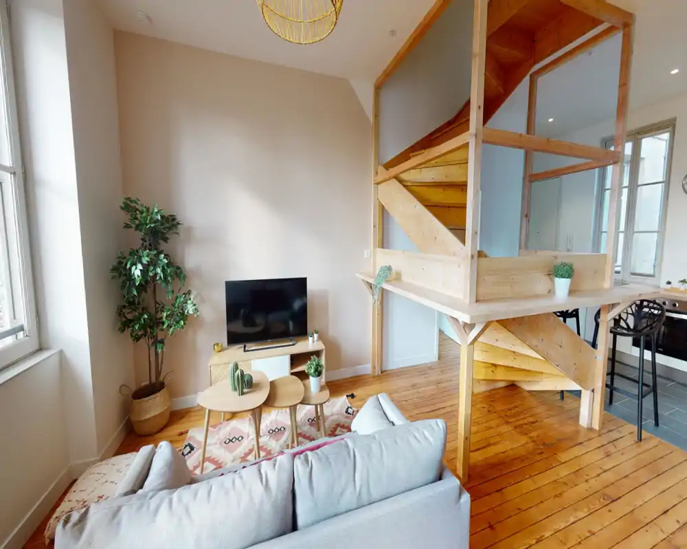 Salon lumineux et chaleureux avec canapé neutre, parquet en bois, tapis décoratif et coin TV compact ; la lumière naturelle des grandes fenêtres et une structure en bois (mezzanine/escalier) apportent du caractère.