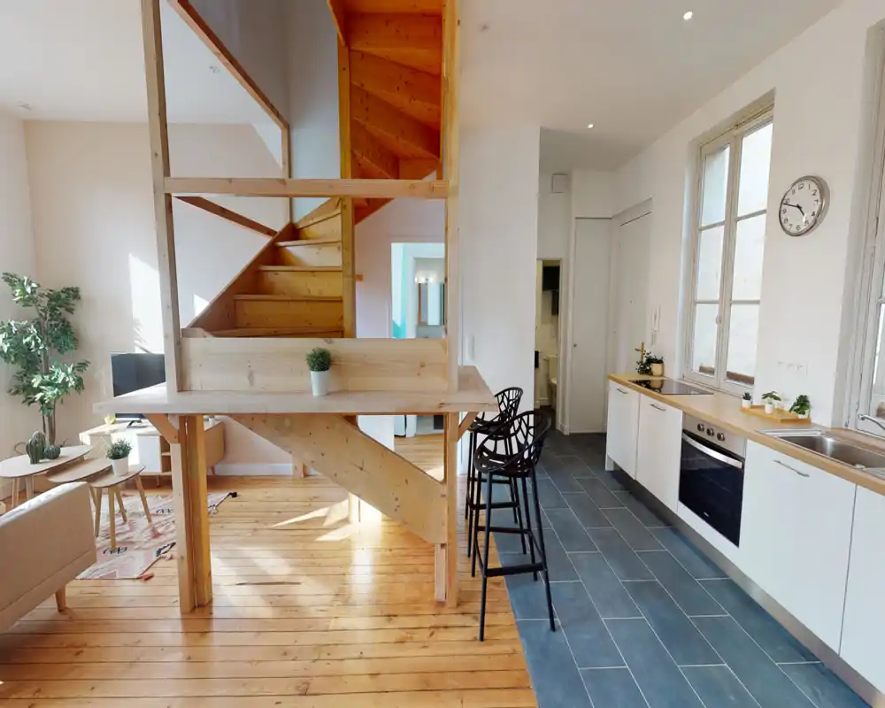 Espace de vie lumineux en open-space avec un escalier en bois et une kitchenette moderne. Lumière naturelle, parquet et plantes décoratives créent une ambiance chaleureuse et aérée, parfaite pour les locations.