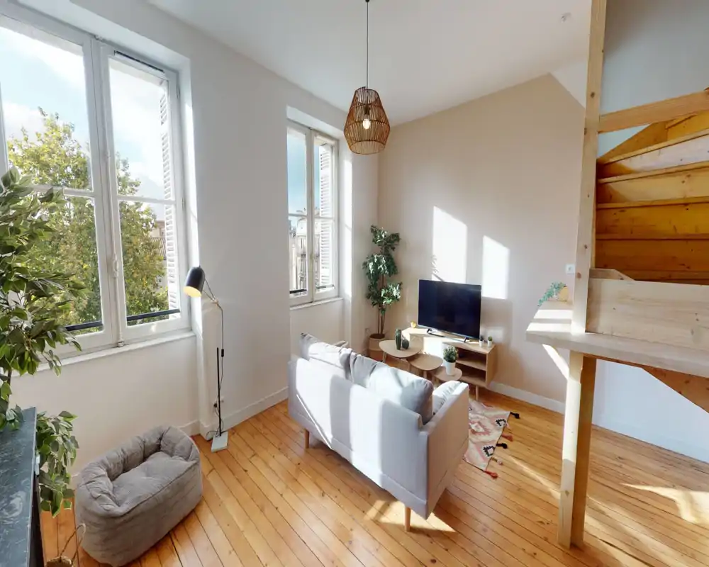 Salon lumineux et cosy avec un canapé compact, un meuble TV et de grandes fenêtres inondant la pièce de lumière naturelle. Parquets chaleureux et plantes pour une ambiance accueillante et soignée.