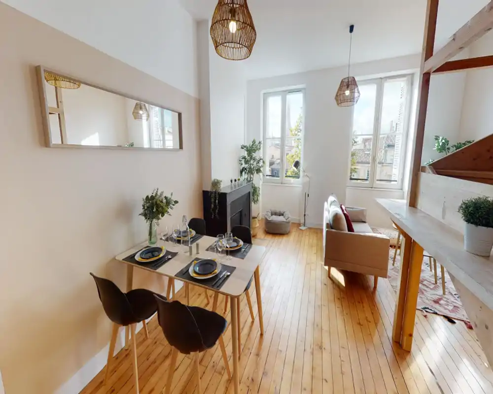 Espace séjour/salle à manger lumineux et décoré avec un sol en bois, grandes fenêtres et suspensions tressées chaleureuses. La pièce est bien mise en scène avec une table dressée pour quatre, un petit canapé et des plantes qui apportent du confort.