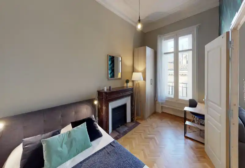 Bright, stylish bedroom with a large upholstered bed, decorative cushions, a classic mantelpiece and a tall window letting in natural light.